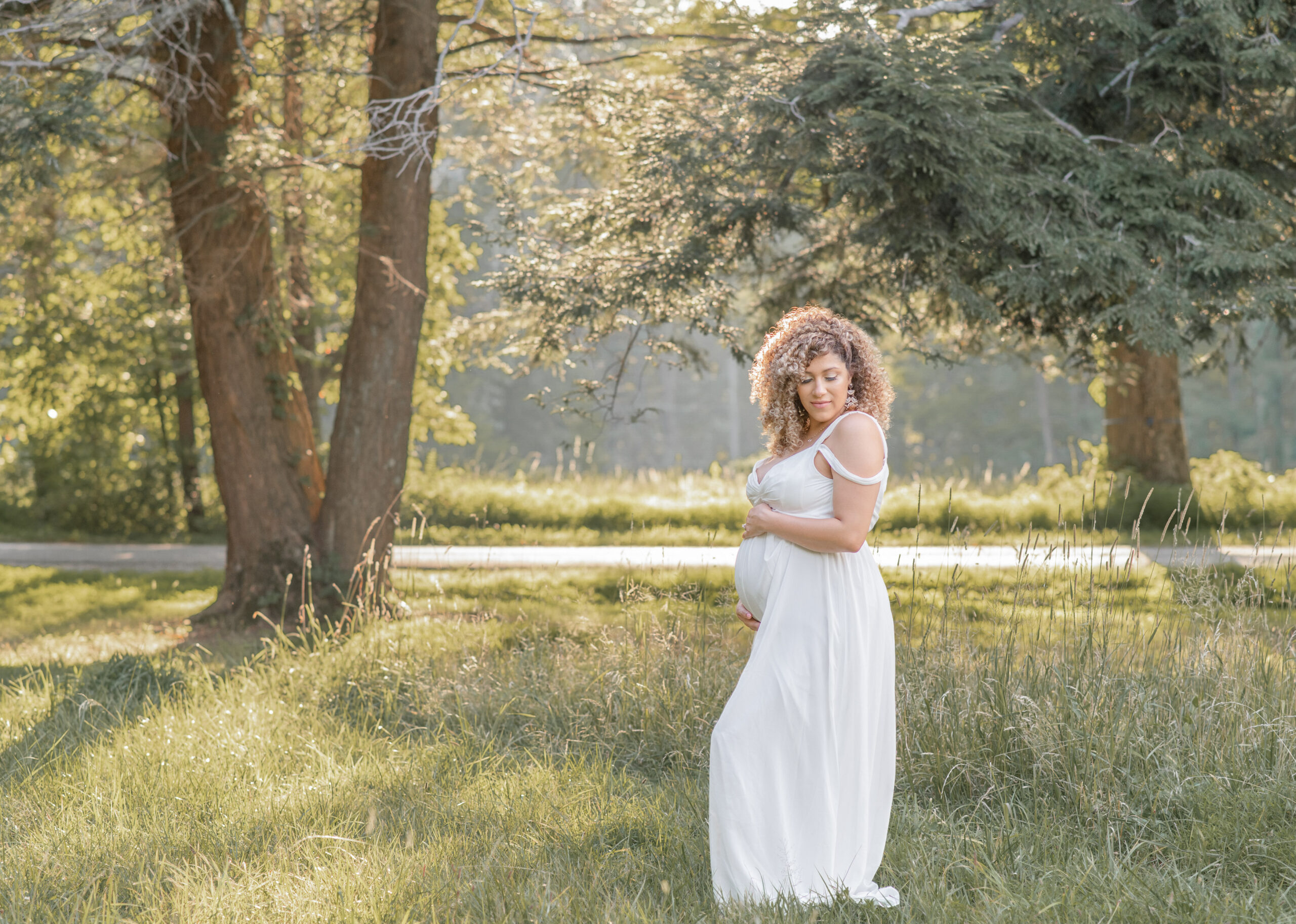 mom to be on field posing holding her belly and wearing a long white dress, beautiful summer day light capture by Elizabeth Klusmann Atlanta maternity photographer