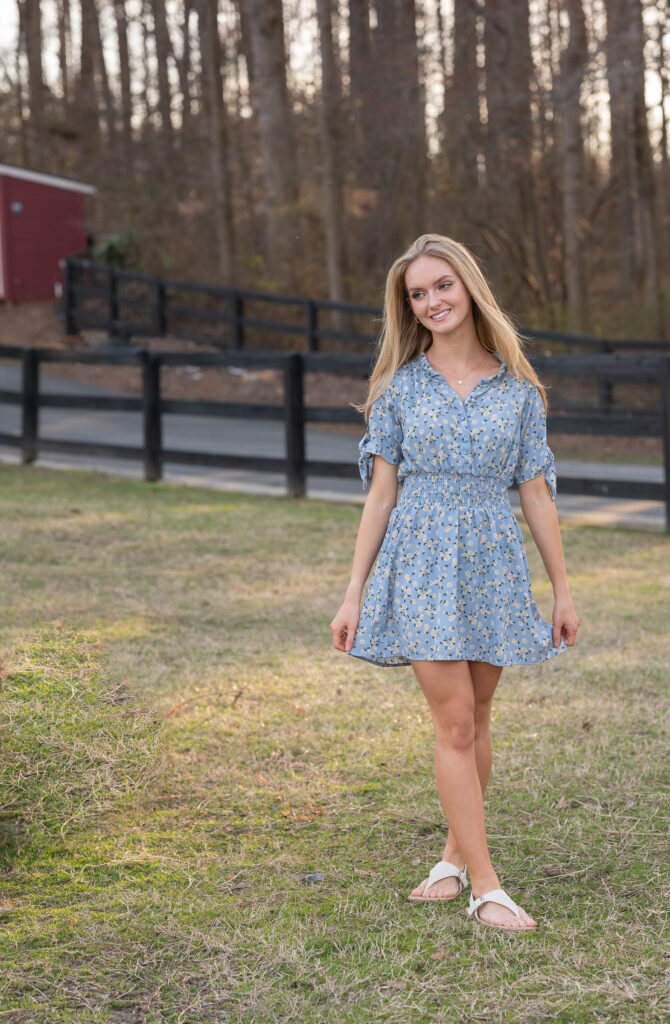 High School senior girl walking and smiling on a field , the sun is on her back and she is wearing a blue dress with small pink flowers on it.