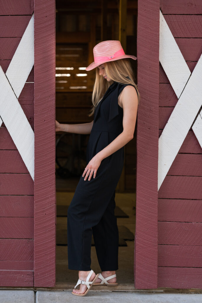 High School senior standing and leaning againts a red barn door, wearing a black outfit and a pink cowboy hat
